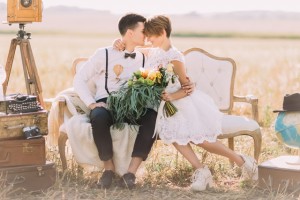 The lovely close-up horizontal portrait of the newlyweds sitting head-to-head on the sofa. The bride is holding the colourful wedding bouquet at the background of the field